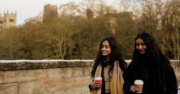 Two women walking on Durham City riverbank holding coffee cups with Durham Cathedral in background.